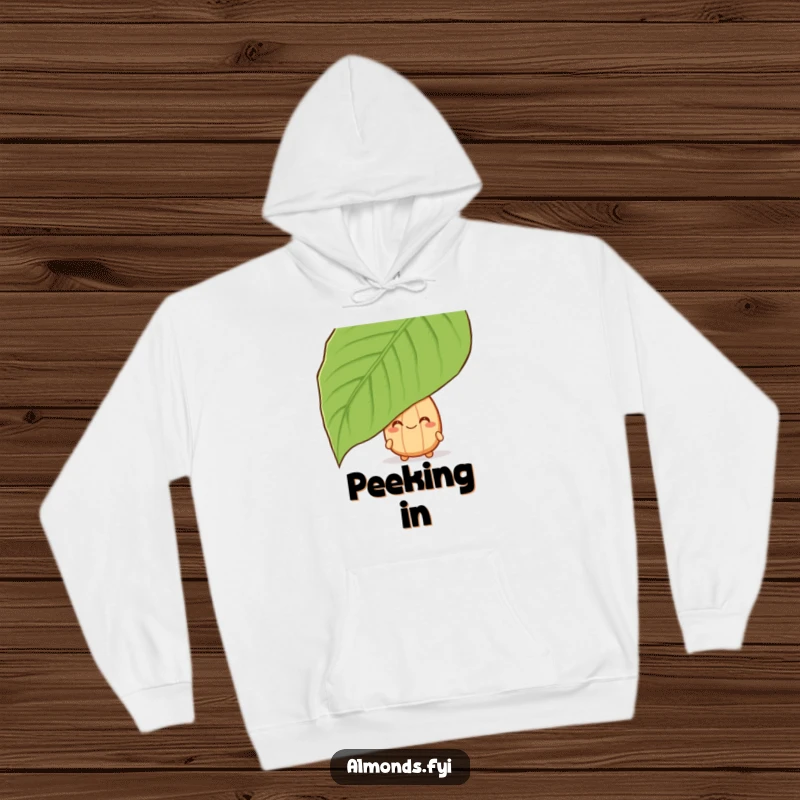 Funny hoodie with a grinning almond character hiding behind a large leaf, offering cozy, playful vibes.